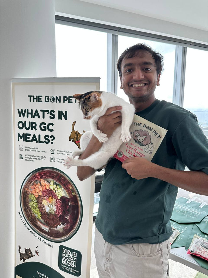 Yash holding a customer's calico cat at a Bon Pet tasting event with the GC meals banner and kraft sample bags - free tastings and personal nutrition answers from the founders